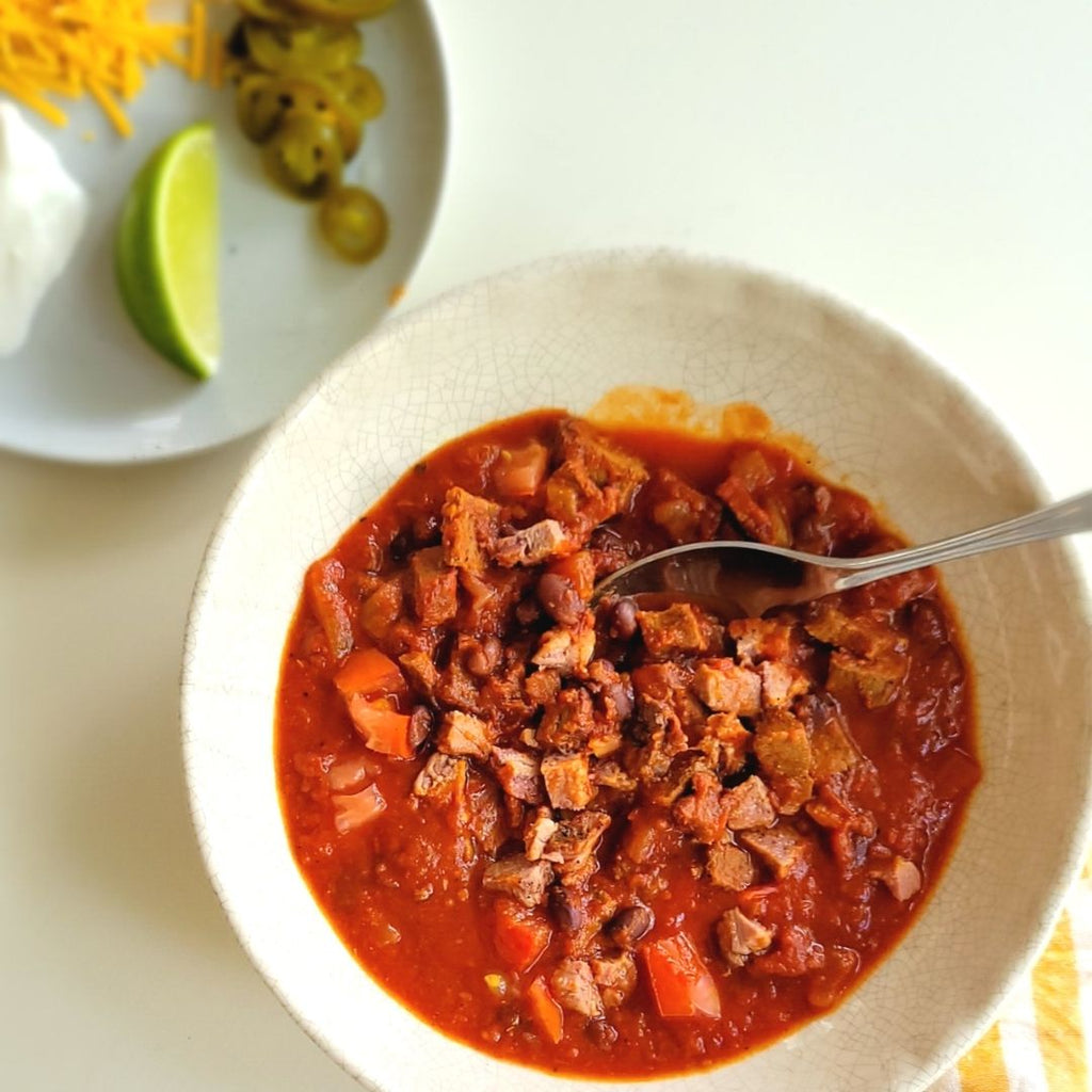 chili in a white bowl with spoon, plate off to side with garnishes.