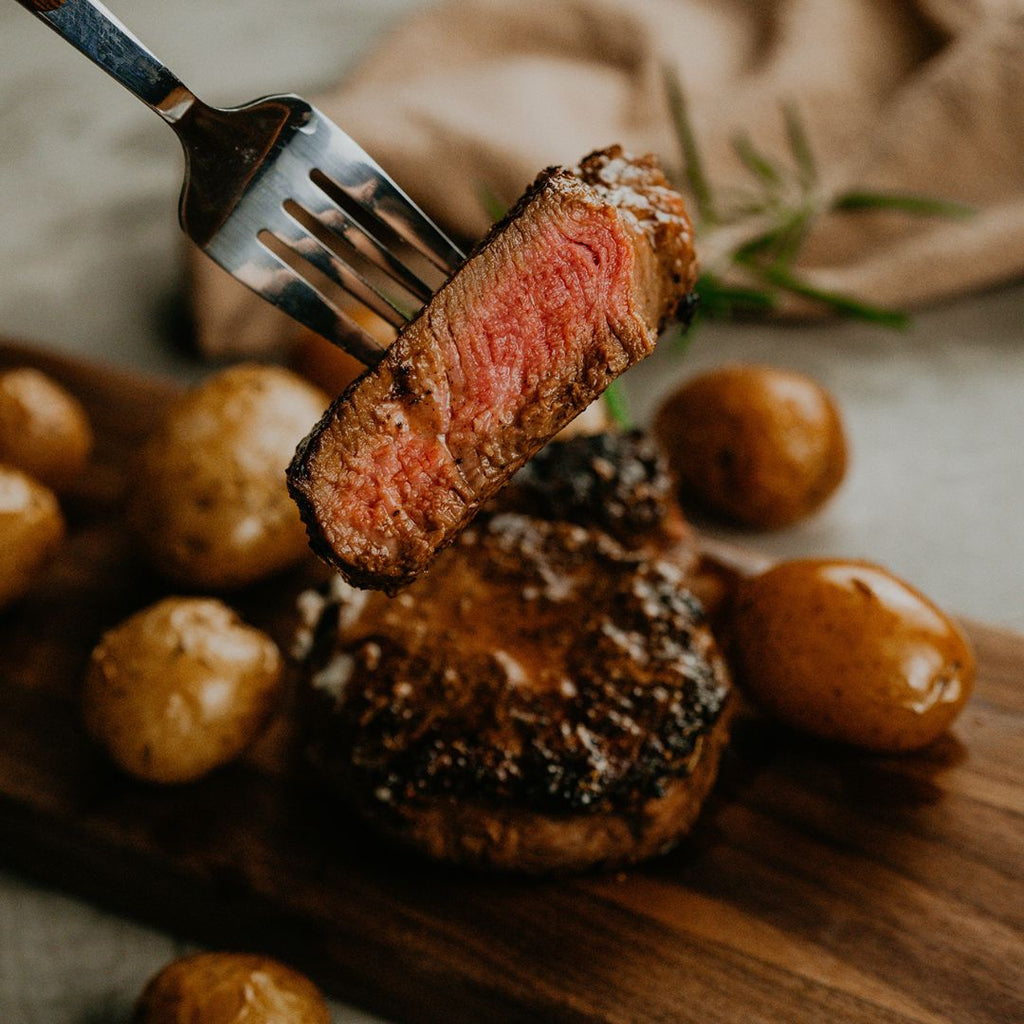 A piece of Bison Filet Mignon steak cooked to rare, held up with a fork like you're about to take a bite. Small, cooked potatoes are in the background on the counter with another cooked steak.