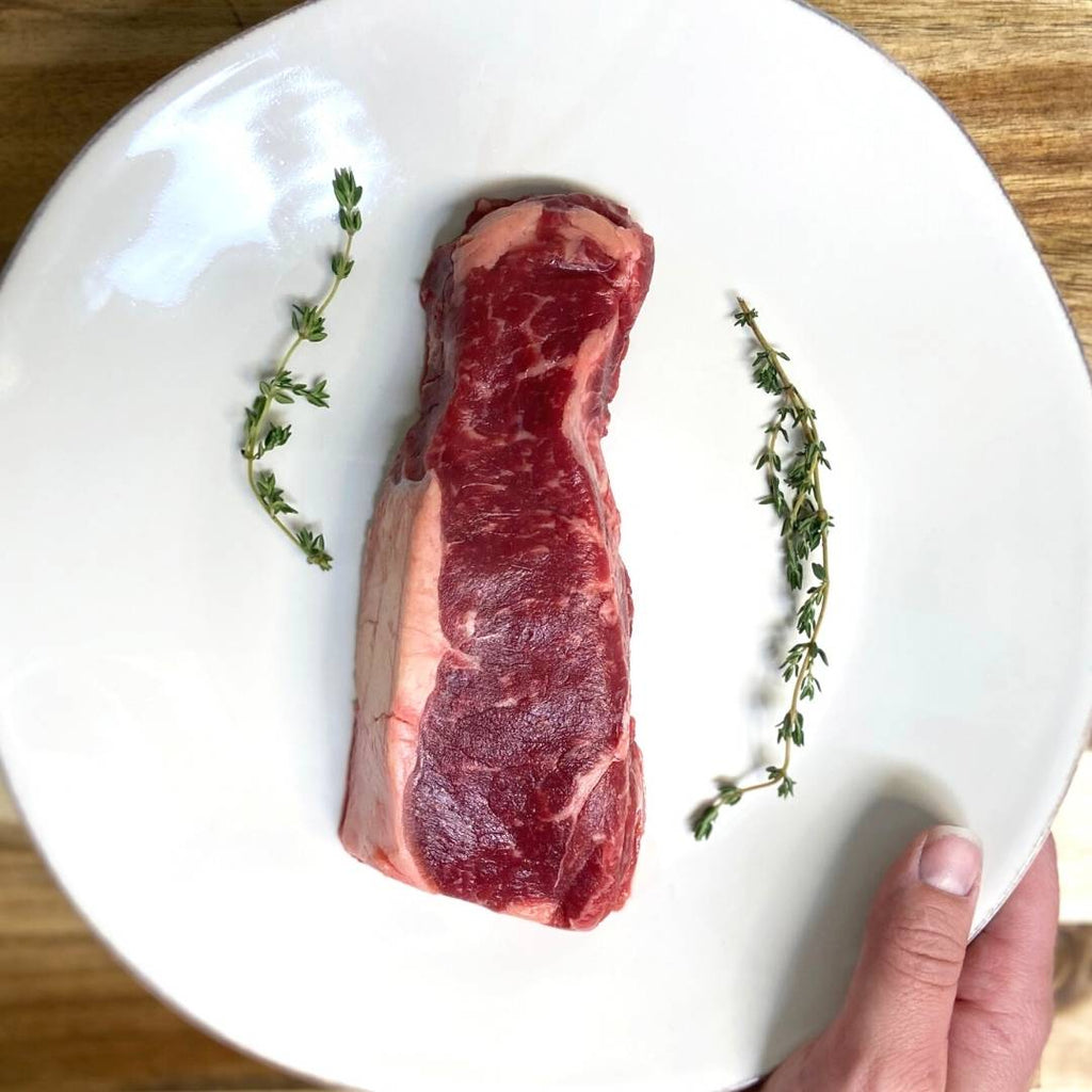 Hand holding a white plate with a raw bison ny strip and sprigs of thyme.