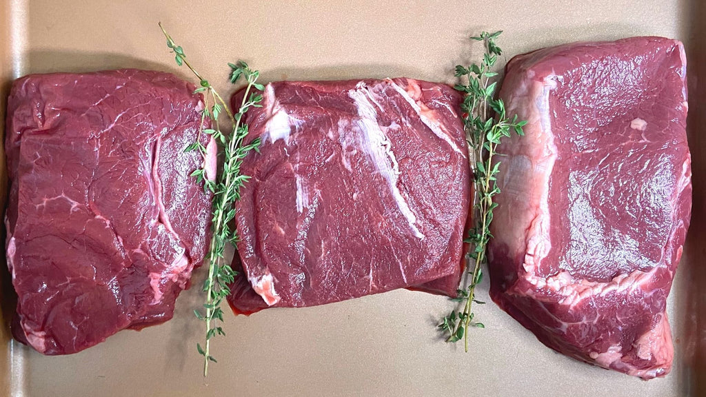 3 raw bison steaks on a tray with thyme sprigs. From left to right: Top Sirloin, Flat Iron, Ribeye.