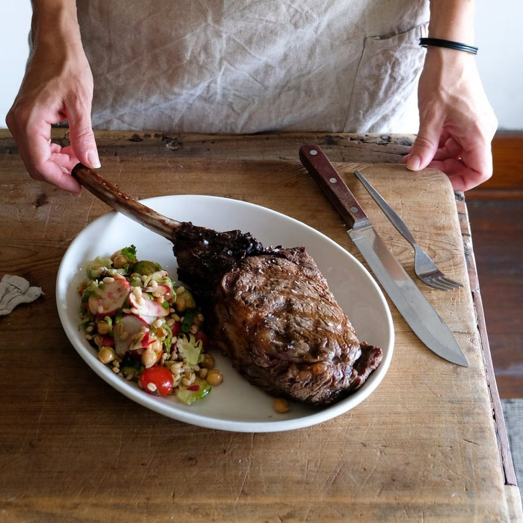 Cooked bison tomahawk steak on white plate with barley salad. On wood counter with knife and fork.