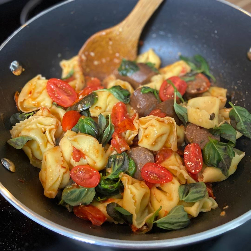 Sliced Bison Hot Italian Sausages in skillet with tortellini pasta, basil and cherry tomatoes.