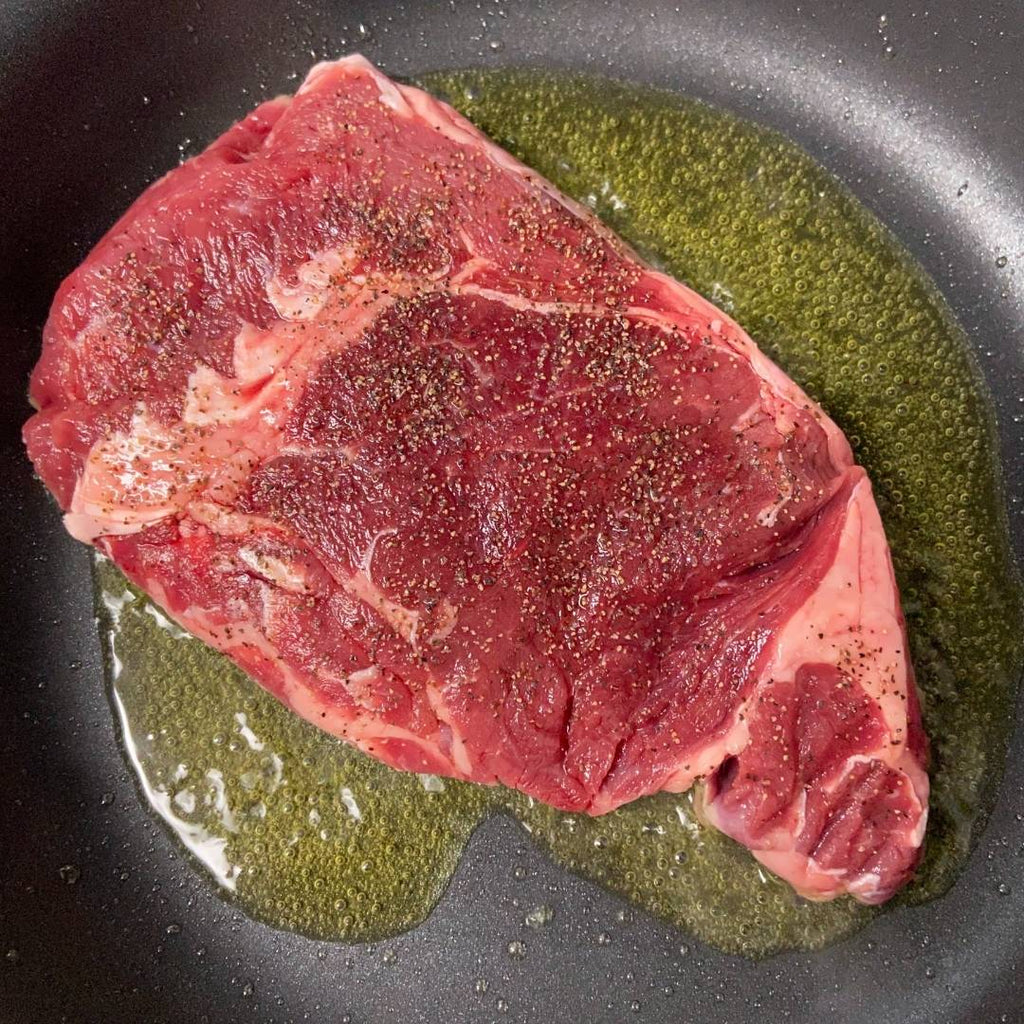 Raw, seasoned, bison ribeye steak in pan with oil.