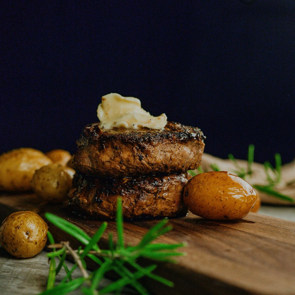 2 cooked Bison Filet Mignon stacked on top of each other with a slab of butter on top. Sitting on a cutting board with small cooked potatoes and rosemary stalks.